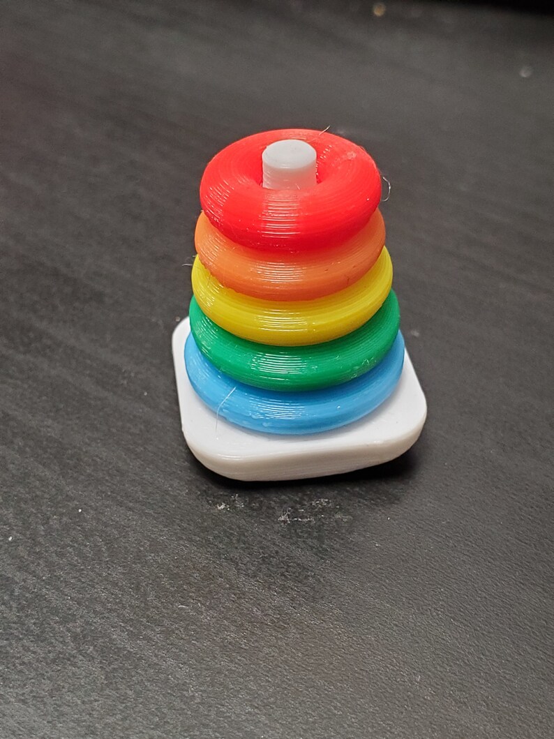 May include: A rainbow-colored stacking toy with a white base. The toy is made of plastic and has six rings that can be stacked on top of each other.