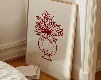 May include: A framed art print featuring a red floral illustration in a vase. The artwork is set against a cream background and framed in a light wood. The print leans against a white wall and wooden floor.