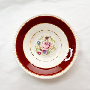 May include: A vintage white bowl with a deep red border and a floral design in the center. The bowl has a gold rim and a beaded edge. The floral design features a pink rose and other colorful flowers.