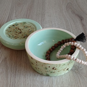 Op de afbeelding: Een lichtgroene keramische sieradenschotel met bruine vlekken, met een deksel en een set houten kralen erin.