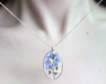 Puede incluir: Un collar dorado con un colgante ovalado. El colgante contiene una flor azul prensada con tallos y hojas verdes. El collar se lleva sobre una blusa negra.