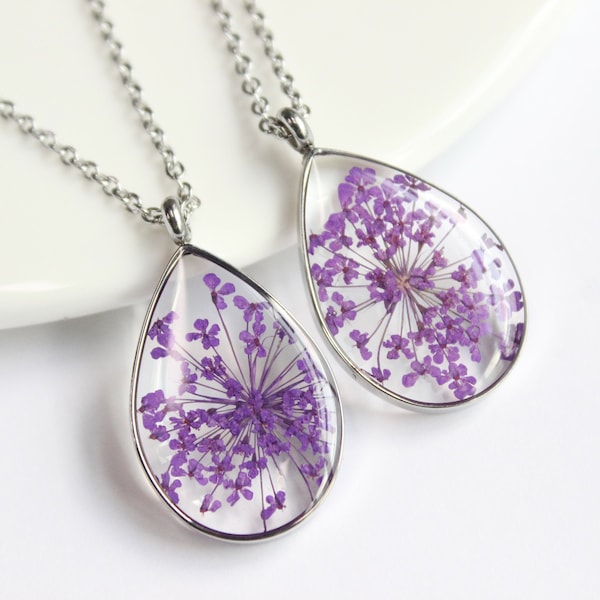 May include: Two teardrop-shaped pendants with silver chains. Each pendant features a clear resin encasing a cluster of small, vibrant purple flowers. The pendants are displayed on a white surface, showcasing the delicate floral details.