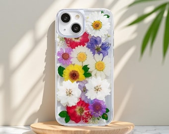 May include: Clear phone case with a variety of colorful dried flowers embedded within. The flowers include white daisies, yellow sunflowers, red and purple blooms, and green leaves. The case is displayed on a wooden stand.