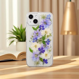 May include: Clear phone case with a floral design. The case features an arrangement of pressed flowers in shades of purple, white, and green. The phone case is on a wooden surface.