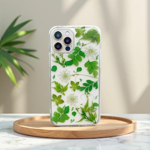 Può includere: Custodia trasparente per telefono con foglie verdi pressate e fiori bianchi. Il design trasparente mette in risalto gli elementi naturali. La custodia è su un vassoio di legno.
