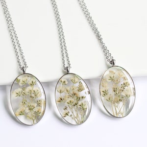 Puede incluir: Tres collares con colgantes ovalados con flores blancas secas encapsuladas en resina transparente. Los colgantes cuelgan de delicadas cadenas plateadas. Los collares se exhiben sobre una superficie blanca, mostrando la belleza natural de las inclusiones florales.