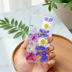 Puede incluir: Funda transparente para teléfono con flores secas incrustadas. La funda presenta una variedad de flores coloridas, incluyendo margaritas blancas con centros amarillos, hortensias moradas y flores azules y moradas. Las flores están dispuestas verticalmente.