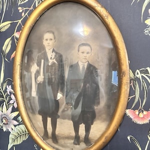 Puede incluir: Fotografía antigua enmarcada en óvalo con dos niños vestidos formalmente. La imagen en tonos sepia está sobre un fondo de papel pintado floral oscuro, con un marco dorado envejecido. Los niños llevan trajes y pantalones cortos.