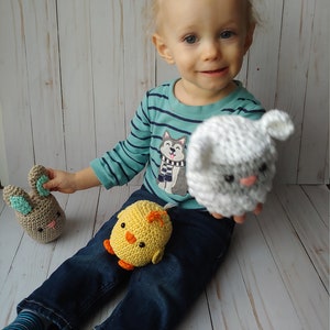 May include: A child is holding three crocheted toys: a tan bunny with blue ears, a yellow chick with orange feet, and a white and gray sheep. The child is wearing a blue striped shirt and blue jeans.