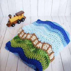 K&ouml;nnte beinhalten: Eine geh&auml;kelte Babydecke in Blau, Gr&uuml;n und Braun. Die Decke hat ein wellenf&ouml;rmiges Muster und einen Rand aus braunem und wei&szlig;em Garn.