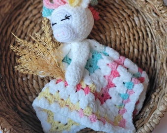 May include: A white crocheted unicorn toy with a rainbow mane and tail, lying on a multi-colored crocheted blanket with yellow, pink, and blue accents.