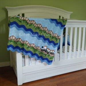 K&ouml;nnte beinhalten: Ein wei&szlig;es Babybett mit einer blauen, gr&uuml;nen und braunen geh&auml;kelten Decke mit einem Wellenmuster und einem Design von Lastwagen. Die Decke ist &uuml;ber das Bett gelegt und h&auml;ngt an der Seite herunter.