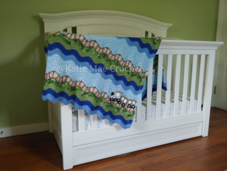 K&ouml;nnte beinhalten: Ein wei&szlig;es Babybett mit einer blauen, gr&uuml;nen und braunen geh&auml;kelten Decke mit einem Wellenmuster und einem Design von Lastwagen. Die Decke ist &uuml;ber das Bett gelegt und h&auml;ngt an der Seite herunter.