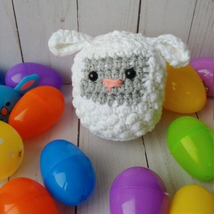 May include: A handmade white crochet lamb with gray accents and black eyes, surrounded by colorful plastic Easter eggs in shades of blue, orange, purple, green, and yellow. The lamb has a pink nose and floppy ears.