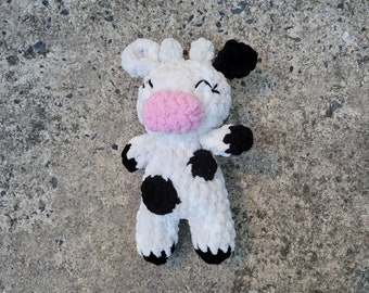 May include: A crocheted plush toy cow with black spots and a pink nose. The cow is lying on a gray concrete surface.