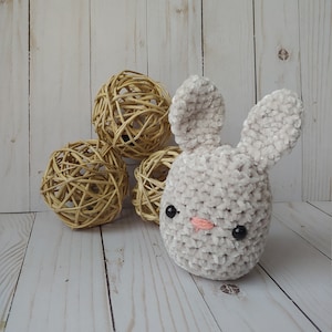 May include: A plush, light gray bunny with black eyes and a pink nose. The bunny is made of soft, textured material. Three woven spheres are in the background. The bunny is a handmade item.