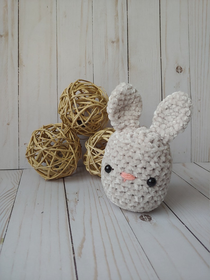 May include: A plush, light gray bunny with black eyes and a pink nose. The bunny is made of soft, textured material. Three woven spheres are in the background. The bunny is a handmade item.