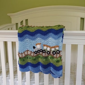 K&ouml;nnte beinhalten: Eine blaue und gr&uuml;ne geh&auml;kelte Babydecke mit einem Zug-Applique. Die Decke liegt &uuml;ber einem wei&szlig;en Babybett.