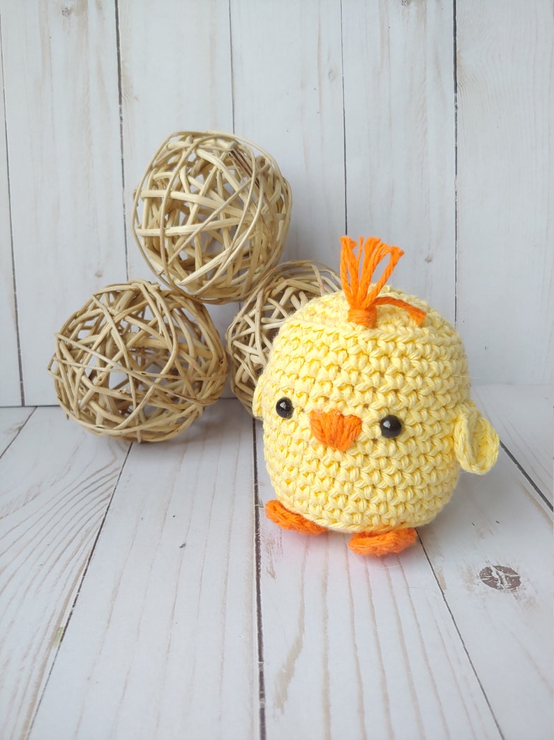 May include: A handmade, yellow crochet chick with orange beak, feet, and head tuft. The chick has black safety eyes and small wings. Behind the chick are three woven rattan spheres, all on a white wooden surface.