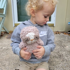 Puede incluir: Un niño pequeño con una sudadera gris con la palabra "NIKE" en la manga sostiene un juguete de peluche marrón y blanco con forma de perezoso. El niño está sentado en un escalón de concreto.