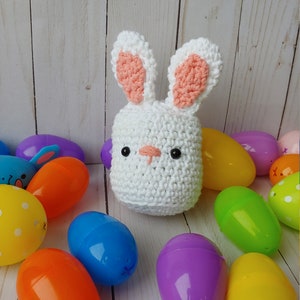 May include: A handmade white crochet bunny with pink inner ears and black safety eyes. The bunny is surrounded by colorful plastic Easter eggs in shades of blue, orange, yellow, green, and purple. The background is a light wood.