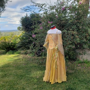 May include: A golden-yellow Renaissance-style dress on a mannequin. The dress features a fitted bodice with embroidered details and long, sheer sleeves. The skirt is flowing, with ribbons. The background includes green grass, trees, and a cloudy sky.