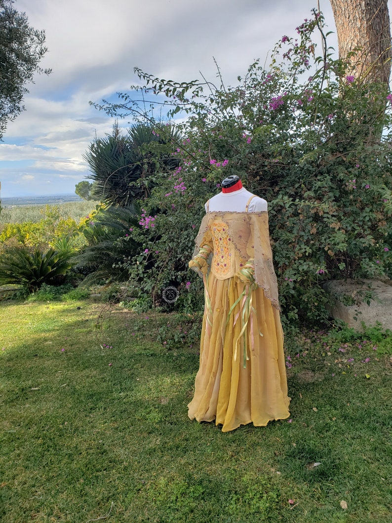 May include: A golden-yellow Renaissance-style dress on a mannequin. The dress features a fitted bodice with embroidered details and long, sheer sleeves. The skirt is flowing, with ribbons. The background includes green grass, trees, and a cloudy sky.