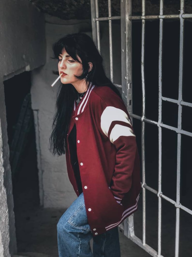 May include: A woman with long black hair wearing a red and white varsity jacket, blue jeans, and a silver necklace. She is standing in a doorway with a metal gate behind her.