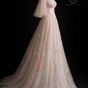May include: A long, blush pink gown with off-the-shoulder sleeves and floral embroidery. The dress has a fitted bodice with a pink ribbon belt and a flowing skirt. The sheer sleeves are flared. The dress is displayed on a mannequin. The label reads 'Raffaella Santoro'.