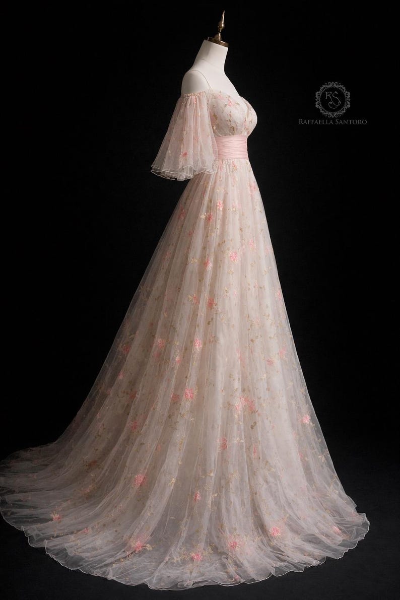 May include: A long, blush pink gown with off-the-shoulder sleeves and floral embroidery. The dress has a fitted bodice with a pink ribbon belt and a flowing skirt. The sheer sleeves are flared. The dress is displayed on a mannequin. The label reads 'Raffaella Santoro'.