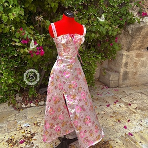 May include: A white satin dress with a pink floral print and a sweetheart neckline. The dress has a fitted bodice and a flowing skirt with a thigh-high slit. The dress is on a mannequin and is surrounded by green foliage.