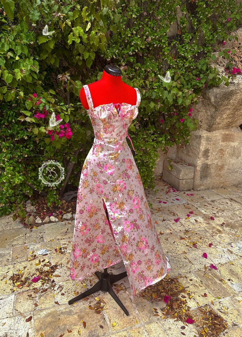 May include: A white satin dress with a pink floral print and a sweetheart neckline. The dress has a fitted bodice and a flowing skirt with a thigh-high slit. The dress is on a mannequin and is surrounded by green foliage.