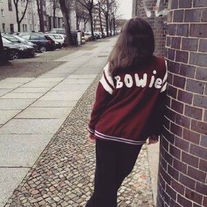 May include: A person wearing a maroon varsity jacket with white and purple stripes and the word "BOWIE" in white lettering on the back. The person is standing on a cobblestone street.
