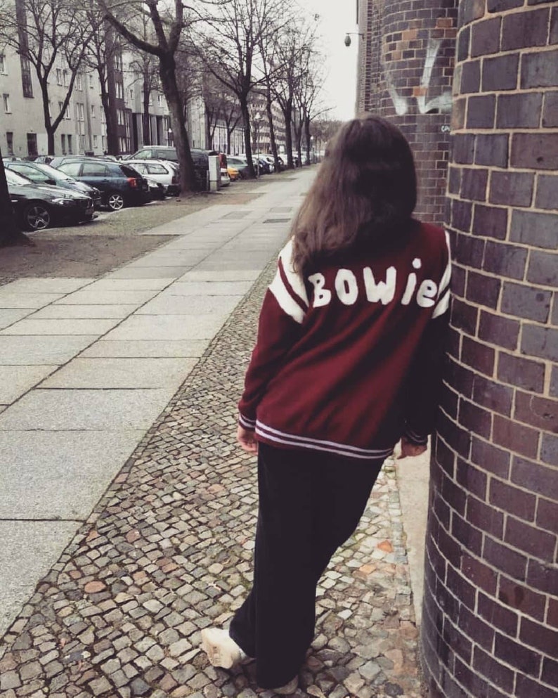 May include: A person wearing a maroon varsity jacket with white and purple stripes and the word "BOWIE" in white lettering on the back. The person is standing on a cobblestone street.