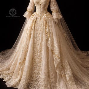 Puede incluir: Vestido de novia color marfil con detalles de encaje. El vestido presenta mangas largas y transparentes, un corpiño ajustado y una falda amplia con múltiples capas. Se aprecian detalles dorados y el vestido tiene una larga cola.