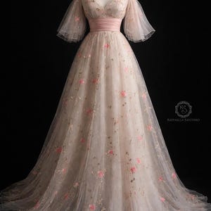 May include: A blush-coloured, off-the-shoulder gown with pink floral embroidery. The dress has a matching pink sash at the waist and sheer, flowing sleeves. The full skirt is layered. The dress is floor-length.