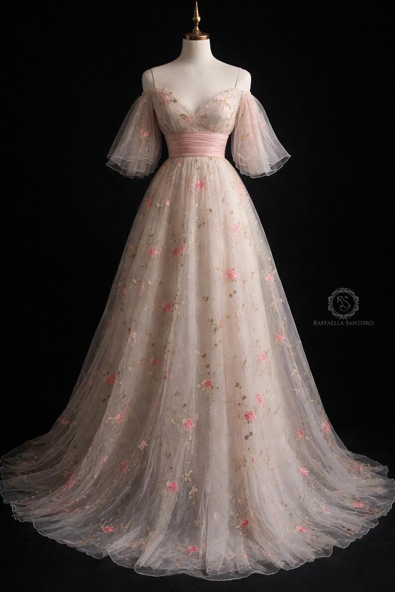 May include: A blush-coloured, off-the-shoulder gown with pink floral embroidery. The dress has a matching pink sash at the waist and sheer, flowing sleeves. The full skirt is layered. The dress is floor-length.