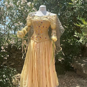 May include: A pale yellow dress with a corset bodice and flowing skirt. The bodice is embroidered with floral designs in shades of red, yellow, and green. The dress has sheer sleeves and is displayed on a mannequin outdoors.
