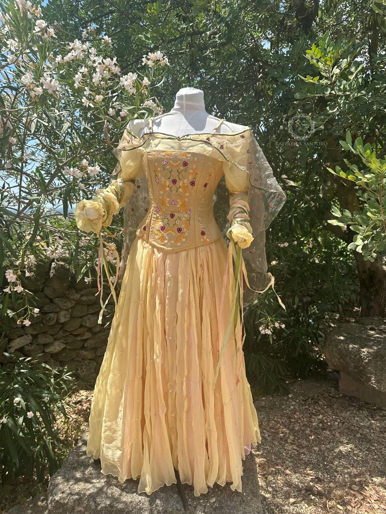 May include: A pale yellow dress with a corset bodice and flowing skirt. The bodice is embroidered with floral designs in shades of red, yellow, and green. The dress has sheer sleeves and is displayed on a mannequin outdoors.