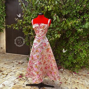May include: A white dress with a pink and orange floral print. The dress has a fitted bodice with a sweetheart neckline and a full skirt. The dress is on a mannequin in front of a green bush.
