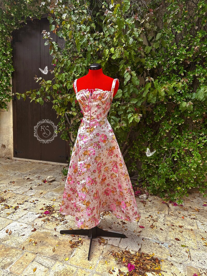 May include: A white dress with a pink and orange floral print. The dress has a fitted bodice with a sweetheart neckline and a full skirt. The dress is on a mannequin in front of a green bush.
