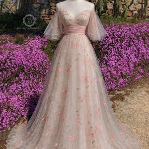May include: A pale pink, off-the-shoulder gown with a flowing skirt. The dress features delicate pink floral embroidery and a pink waistband. The bodice has a sweetheart neckline and sheer, flutter sleeves. The dress is displayed on a mannequin in front of a stone wall and purple flowers.