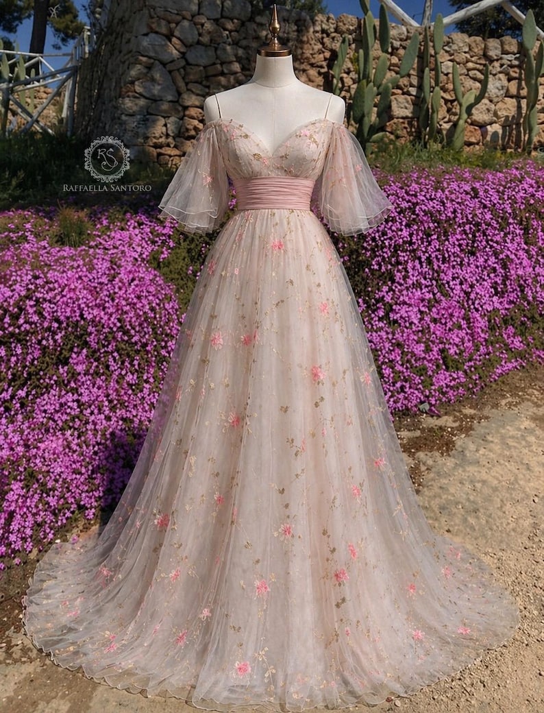May include: A pale pink, off-the-shoulder gown with a flowing skirt. The dress features delicate pink floral embroidery and a pink waistband. The bodice has a sweetheart neckline and sheer, flutter sleeves. The dress is displayed on a mannequin in front of a stone wall and purple flowers.