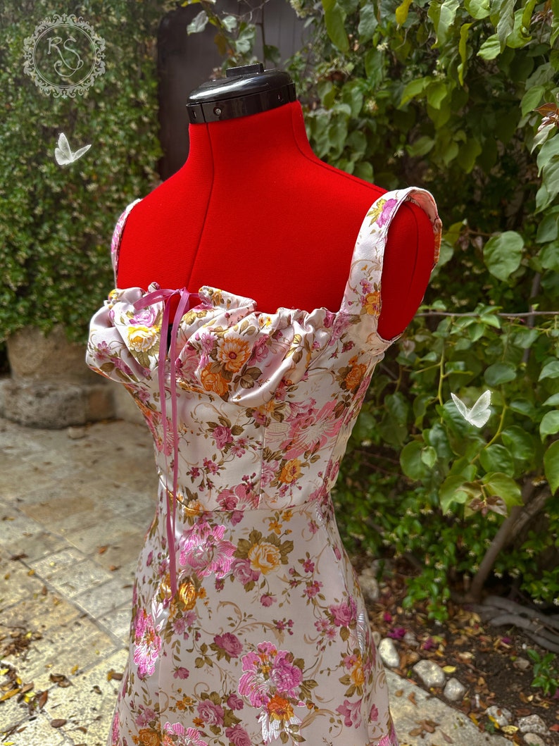 May include: A white satin dress with a floral print and pink straps. The dress has a gathered neckline and a fitted bodice.