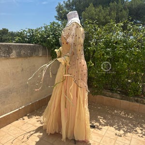 May include: A flowing, layered dress in shades of pale yellow and pink, with a sheer overlay adorned with floral embroidery. The dress features long sleeves and a matching head covering. The dress is displayed on a mannequin outdoors.