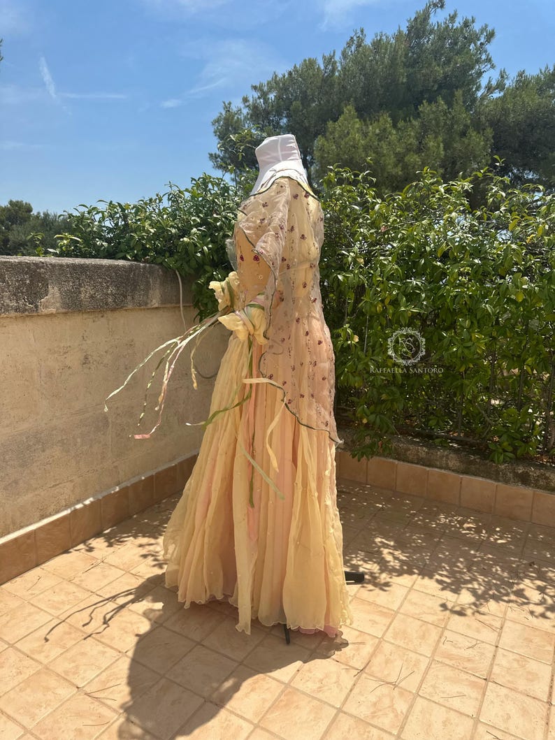 May include: A flowing, layered dress in shades of pale yellow and pink, with a sheer overlay adorned with floral embroidery. The dress features long sleeves and a matching head covering. The dress is displayed on a mannequin outdoors.