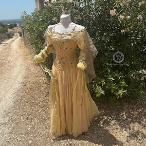 May include: A light yellow dress with a floral embroidered bodice and long sleeves. The dress features a flowing skirt and a sheer cape. The dress is displayed on a mannequin outdoors, with a path and greenery in the background.