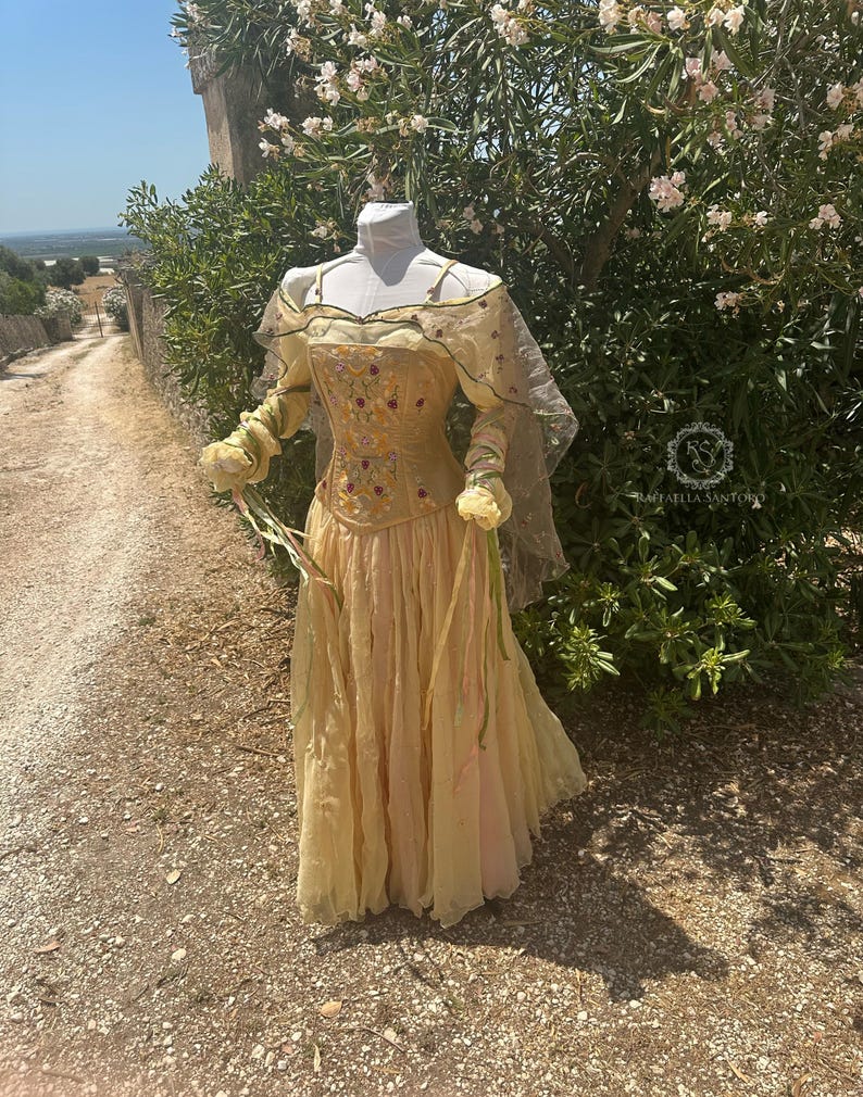 May include: A light yellow dress with a floral embroidered bodice and long sleeves. The dress features a flowing skirt and a sheer cape. The dress is displayed on a mannequin outdoors, with a path and greenery in the background.