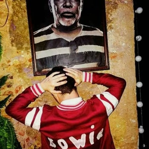 May include: A person wearing a red and white striped bomber jacket with the word "BOWIE" embroidered on the back. The person is standing in front of a framed picture of a man with a white beard.