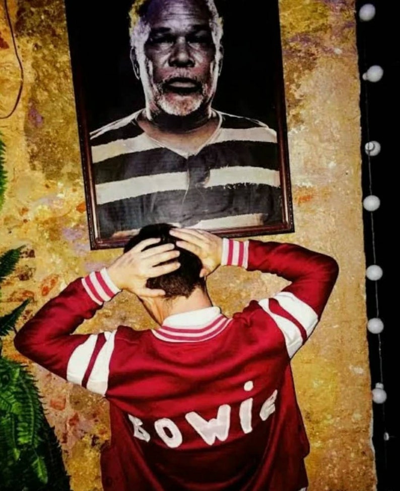 May include: A person wearing a red and white striped bomber jacket with the word "BOWIE" embroidered on the back. The person is standing in front of a framed picture of a man with a white beard.